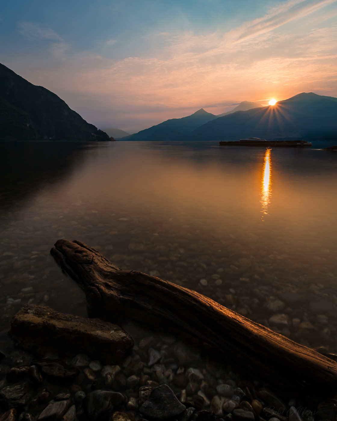 Long exposure of sunrise in Menaggio, Lake Como, featuring a log in the foreground, sunburst behind mountain silhouettes, and warm orange light spreading across the water under a soft blue morning sky.