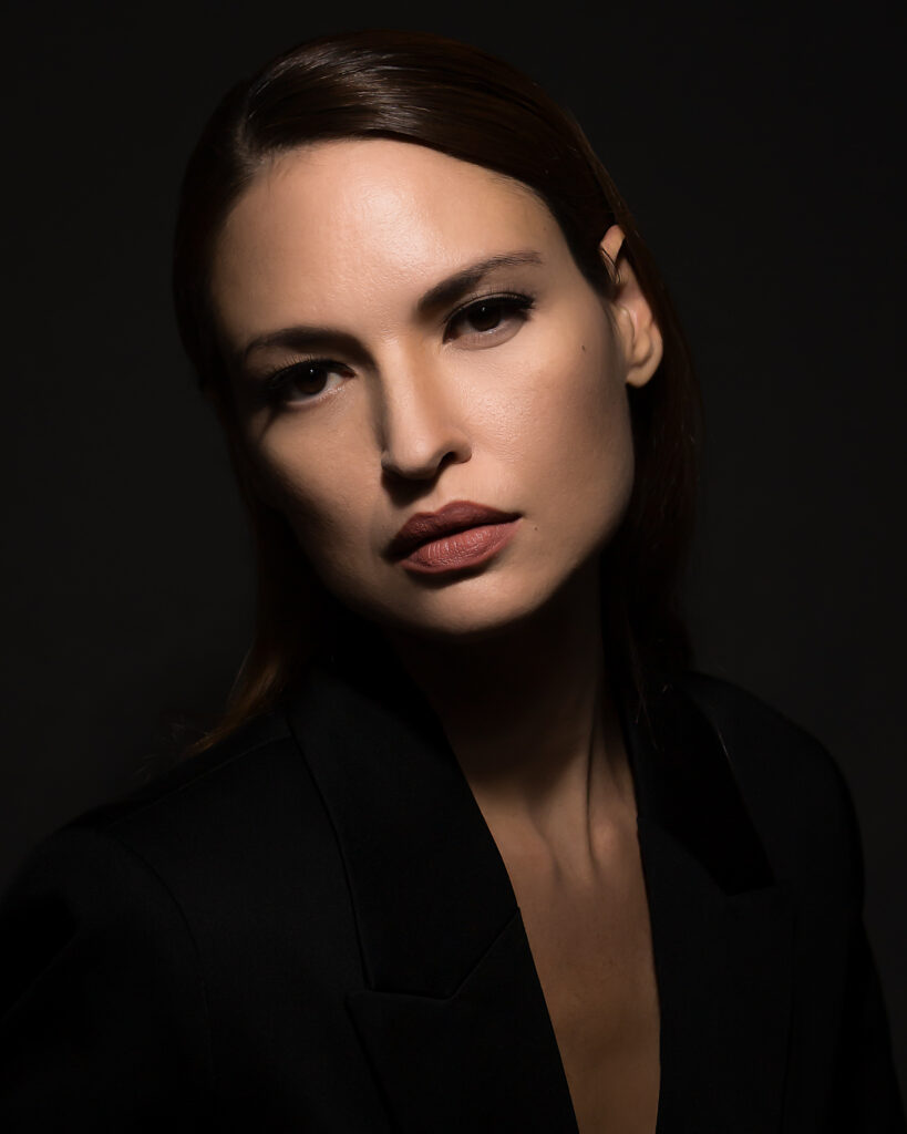 Model Bibi Alfonso in a black blazer, photographed in dramatic directional lighting against a dark background.