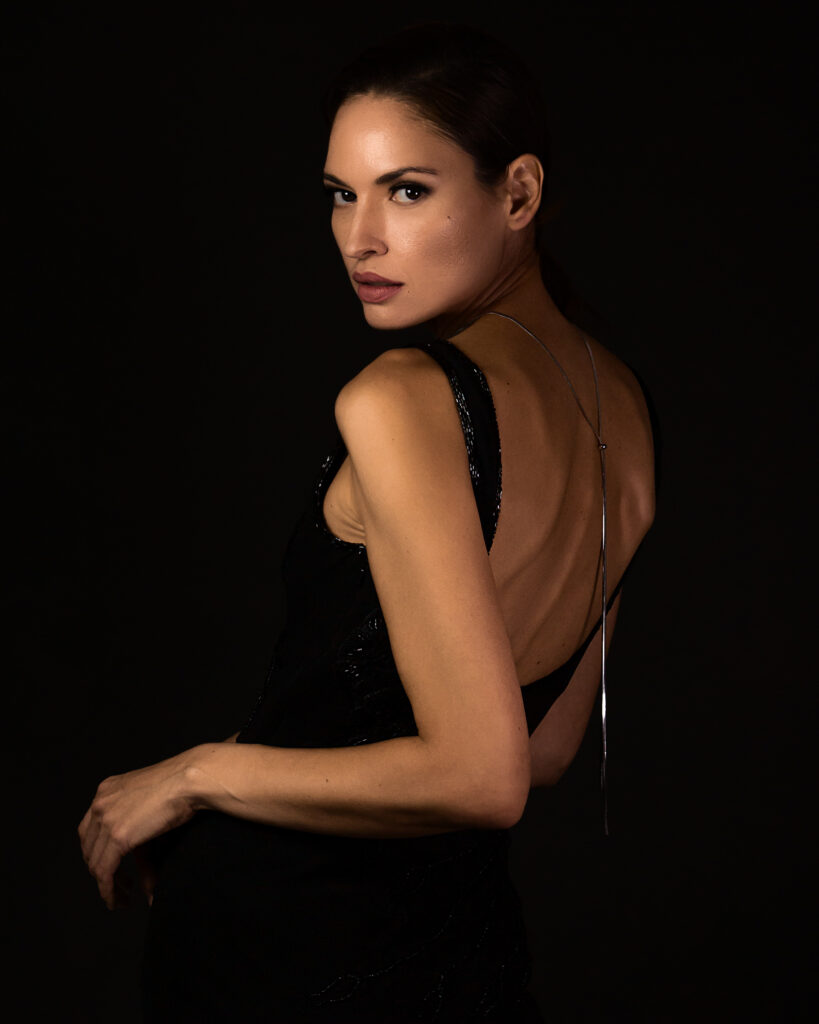 Model Bibi Alfonso posing in a black evening dress with an open back against a dark background.