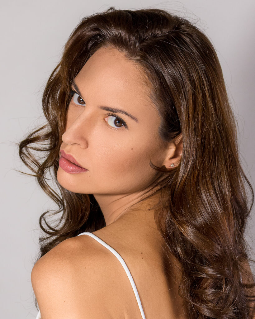 Model Bibi Alfonso looking over her shoulder in a white top, with long brown hair against a light background.