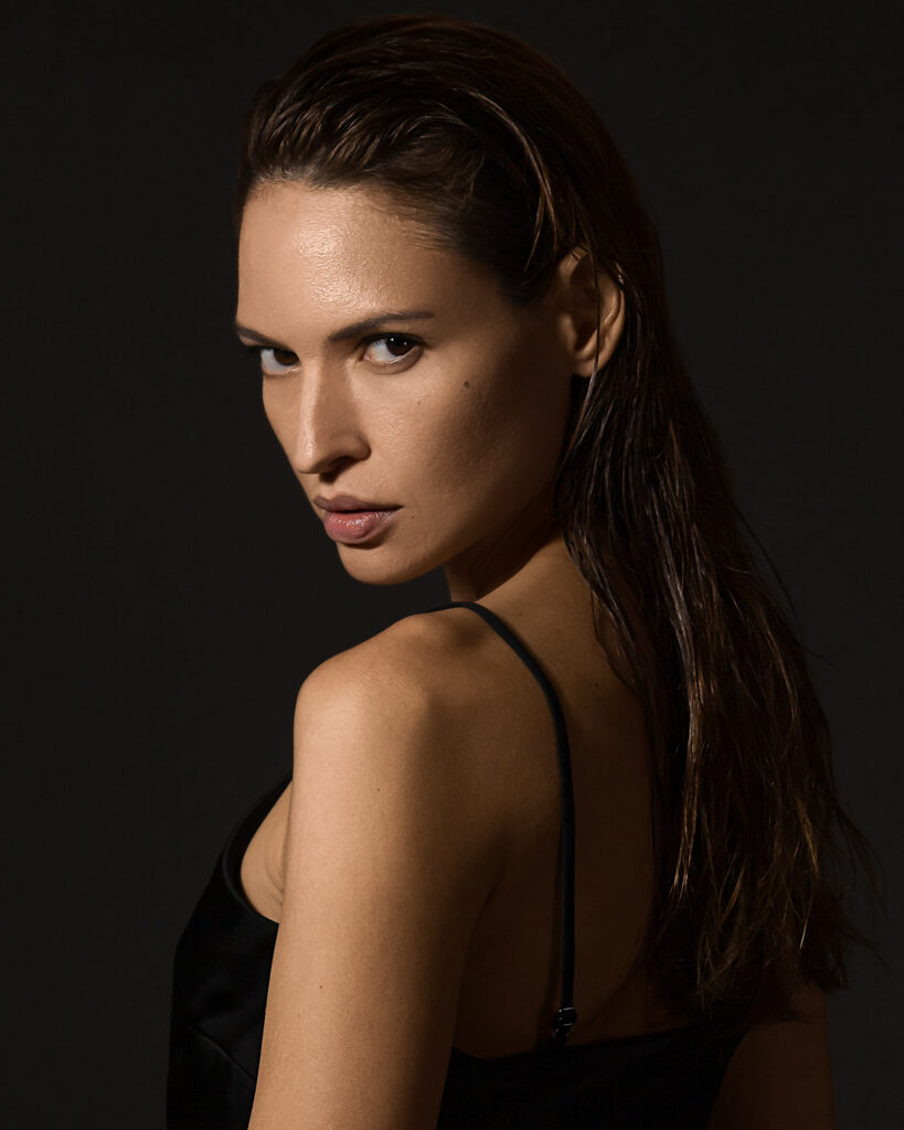 Model Bibi Alfonso looking over her shoulder in a black dress against a dark background.